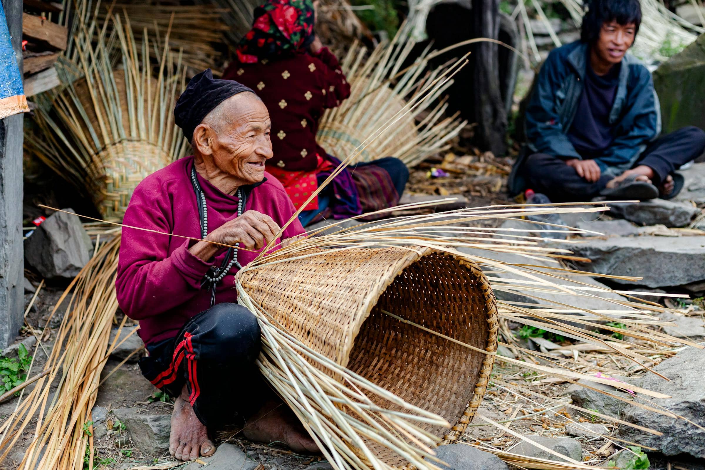 An elder Nepali artisan weaving a traditional bamboo basket by hand in a village courtyard near Jyamrung.