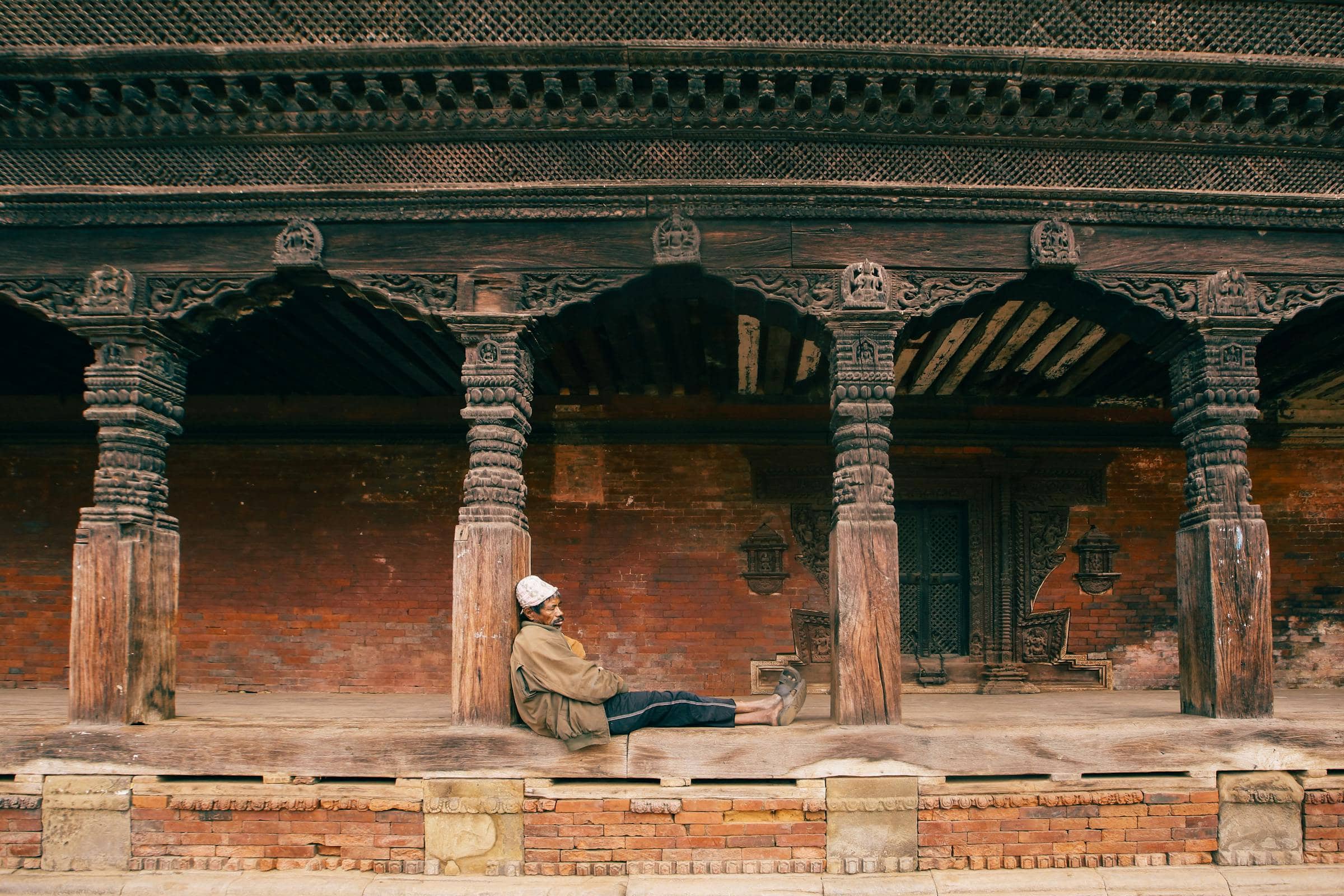A traveler resting against a carved wooden pillar in a Patan courtyard — the practice of arrival.