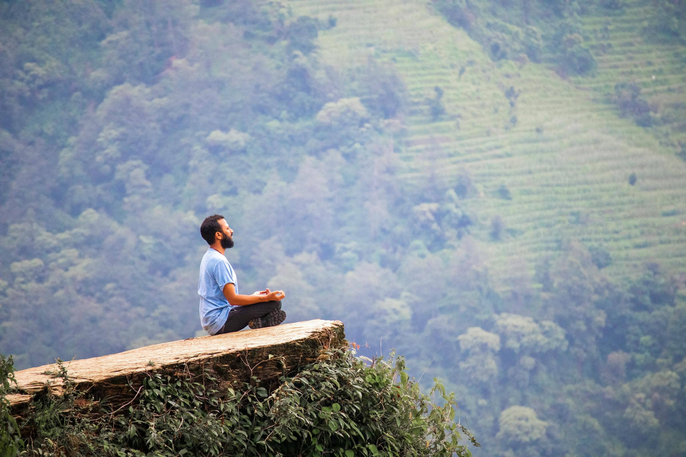 A solitary practitioner in seated meditation on a wooden ledge above the terraced hills of Pokhara at dawn.